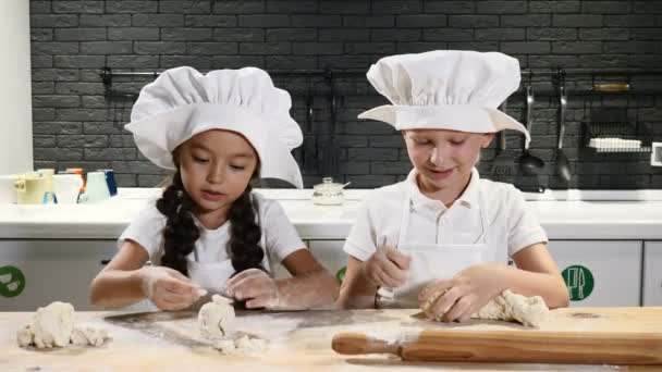 Kinder kochen in der Küche. Zwei kleine Kinder in Kochmützen rollen lächelnd Teig. 4k — Stockvideo