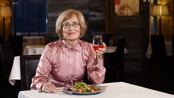 Aantrekkelijke senior vrouw in haar late 70s zit in een restaurant met een glas vruchten drank in de hand. Lachende en blije. Gelukkige oude dag. 4k — Stockvideo