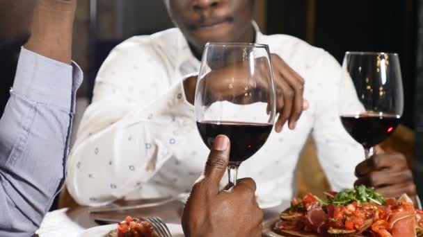Due amici africani irriconoscibili al ristorante seduti a tavola davanti a un bicchiere di vino. Mano africana maschile che regge un bicchiere di vino. 4k — Video Stock
