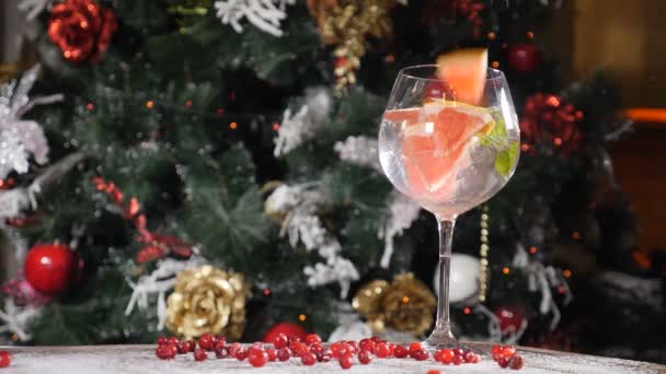 New Year and Merry Christmas table concept. Feast cocktail in a glass with a straw in it with a fur tree in background. A slice of orange is falling into a cocktail splashing. hd — Stock Video