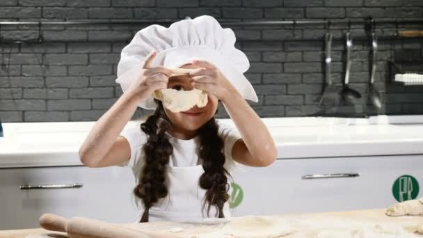 Menina bonito jogando na cozinha doméstica. Massa, farinha e rolo de pino na mesa. As crianças agem como adultos. Menina engraçada em chapéu de chef e avental cozinhar biscoitos apreciando o processo. 4k — Vídeo de Stock