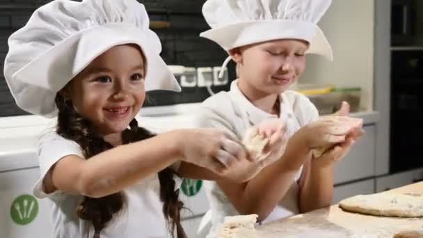 Les petits enfants jouent aux chefs avec la pâte, le rouleau à pâtisserie et la farine. 5-6 ans travaillant ensemble dans des tabliers et des chapeaux de chef dans la cuisine domestique. 4k — Video