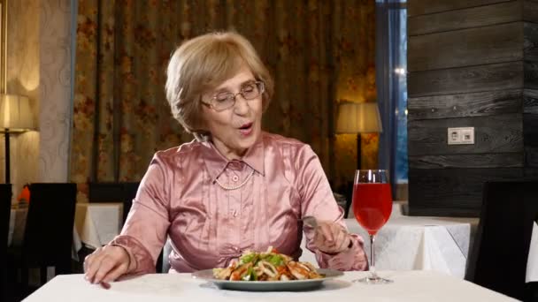 Une femme à la retraite est assise au restaurant avec une salade de légumes et un verre de boisson aux fruits frais sur la table. Un mannequin dans la soixantaine. 4k — Video