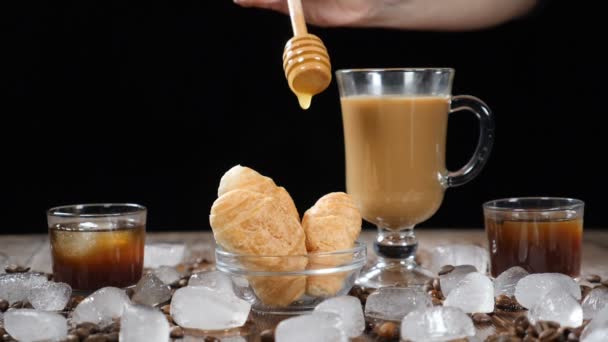 Koffiehuis met heerlijke croissants op de glasplaat. Warm schakelt u vloeibare honing gieten neer op dessert in slow motion. Kopje aromatische latte op de achtergrond. Ijsblokjes, koffiebonen op houten bord — Stockvideo