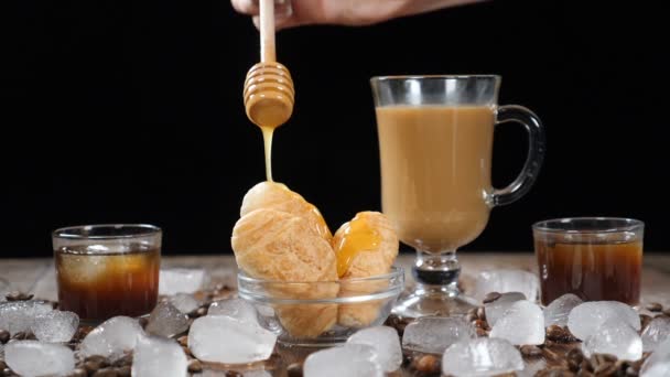 Kaffeehaus mit leckeren Croissants auf Glasplatte. heißer flüssiger Honig, der sich in Zeitlupe auf das Dessert ergießt. Tasse aromatischen Latte auf dem Hintergrund. Eiswürfel, Kaffeebohnen auf Holzbrett — Stockvideo