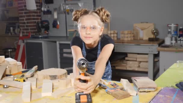 Porträt eines lustigen 10-jährigen Mädchens in einer Holzschreinerei, das eine elektronische Bohrmaschine hält und vor der Kamera posiert. Wenig Bauherrenkonzept. hd — Stockvideo