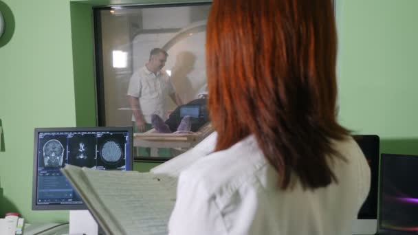 Especialista em radiologia feminina em casaco branco assistindo paciente recebendo procedimento de TC MRI e twriting resultados médicos durante o exame de raios-X, tiro steadycam. Conceito de tomografia computadorizada. vista de fora — Vídeo de Stock