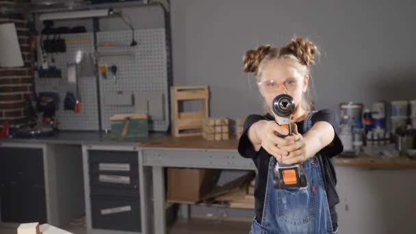 Portrait of 10 year old girl in wood carpentry holding an electronic drill, posing at camera. Little builder concept. hd — Stock Video
