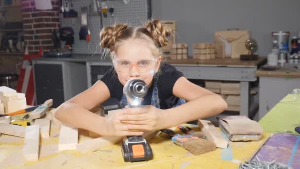 Porträt eines lustigen 10-jährigen Mädchens in einer Holzschreinerei, das eine elektronische Bohrmaschine hält und vor der Kamera posiert. Wenig Bauherrenkonzept. hd — Stockvideo