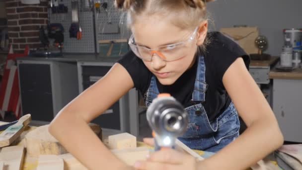 Porträt eines lustigen 10-jährigen Mädchens in einer Holzschreinerei, das eine elektronische Bohrmaschine hält und vor der Kamera posiert. Wenig Bauherrenkonzept. hd — Stockvideo