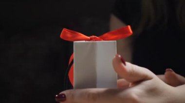 Karanlık görüntü. Parmaklar gösterir kağıt hediye kutusu kırmızı kurdele ve bir yay ile kapatın. hediye kutusu kadın olarak tutan bir hediye açılış yay-knot siyah bir zemin üzerine bağcıklı. HD