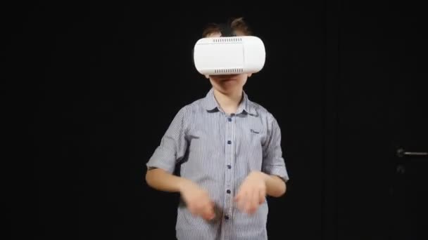 Beau jeune garçon d'âge scolaire en lunettes VR avec un fond noir. Petit garçon dans un casque de réalité virtuelle debout dans une pièce sombre et regardant autour de lui avec étonnement. Gros plan. garçon regarde autour — Video