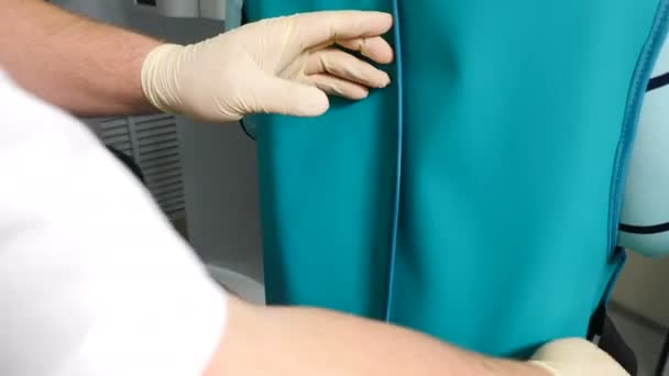 Antes do procedimento de raio-x na clínica moderna. Foto retrovisada de um médico fixando o cinto no avental de chumbo. O dentista coloca o cobertor de raios-X no paciente. Conceito de saúde. 4k — Vídeo de Stock