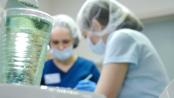 Tiro de vidrio de plástico con agua en el primer plan y dentista borroso y asistente haciendo procedimiento dental en el fondo. Primer plano de las manos del dentista y la joven con cofferdam en la boca. Doctor. — Vídeo de stock