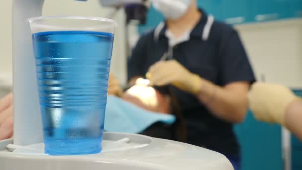 첫 번째 계획 및 흐리게 치과 의사와 보조 배경에 치과 절차를 만들기에 파란색 액체 플라스틱 유리의 샷. 치과 의사 손 및 치과 자에서 젊은 여자의 근접 촬영 샷. 의사 — 비디오
