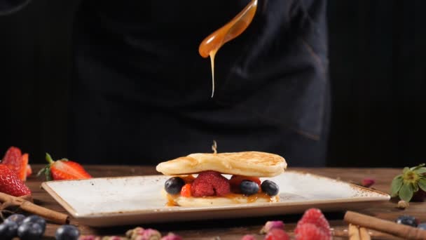 Arte culinario. Chef esparciendo caramelo en la parte superior de la pila de panqueques esponjosos frescos decorados con bayas del bosque. Panqueques estilo americano mullidos veganos sanos caseros con frutas frescas. Verter miel en — Vídeo de stock