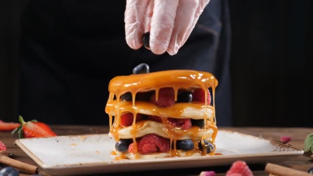 Close-up van chef-kok handen gieten organische bramen op heerlijke biologische pannenkoeken voor ontbijt thuis of in een restaurant. Voedsel video. Op zwarte achtergrond. HD. Slow motion — Stockvideo