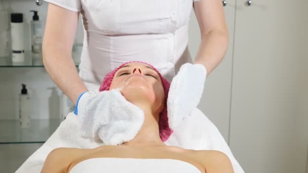 Concepto de salud y belleza. Beautician elimina la máscara cosmética facial de la cara del cliente femenino. Primer plano de las manos con guantes de toalla tibios limpiando la cara femenina de mediana edad. hd — Vídeo de stock