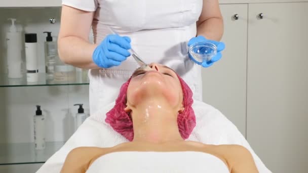 Skönhets klinik. Ung kvinna får professionell ansikts behandling. Kosmetolog sprider mask på kvinnligt ansikte med en borste. renovering av ansiktet. Hd — Stockvideo