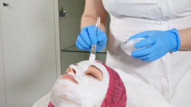 Yüze beyaz anti-aging maskesi uygulayan güzellik uzmanı. Güzellik konsepti. Spa merkezinde 30'lu yaşlarda genç bir kadın yüz germe işlemi yaptırıyor. Yakından kapatın. Hd