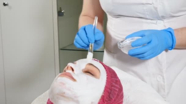顔に白いアンチエイジングマスクを施した美容師。美容コンセプト。スパセンターの30代の若い女性は、顔の持ち上げ手順を取得します。クローズアップ。Hd — ストック動画