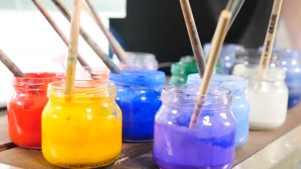 Bright glass jars with aquarelle paint inside. Ebru art. Close up. Paint brushes in jars. Many different acrylic paints in jars. female hand holds paint brush. Paint drop. 4k — Stock Video