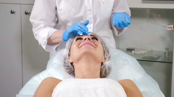 Conceito de esteticista. Cosmetician que cobre a cara feminina com a nata anestésica antes do procedimento de beleza. Mulher bonita nova recebendo máscara nutritiva facial por esteticista no salão de spa. 4k — Vídeo de Stock