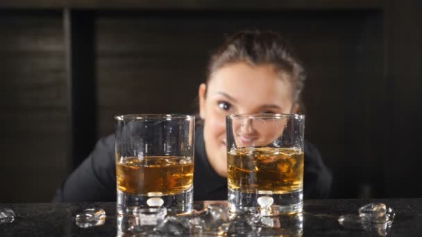バーカウンターの後ろに立つウイスキーグラスを通してカメラを見ている幸せな笑顔の女性バーテンダーの肖像画。眼鏡が集中している。Hd — ストック動画