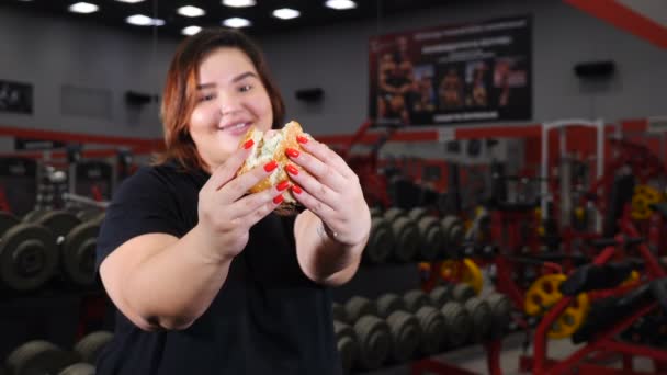 Portrait of obese woman in gym eating hamburger smiling and looking at camera. Food temptation. Overweight concept. Sport with unhealthy food. 4k — Stock Video