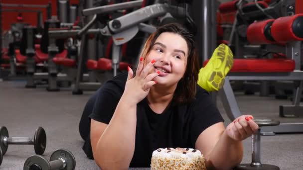 Mladá, příjemná obézská žena ležící v tělocvičně a s lahodným koláčem. Koncept willpower. Autentický fitness životní styl a jídlo. 4k — Stock video