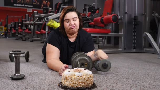 Deporte con alimentos poco saludables. Joven mujer gorda más tamaño guarda las barras y se lleva un pastel grande. Mujer joven que parece confundida y culpable en el gimnasio. comida chatarra y malsana. 4k — Vídeos de Stock