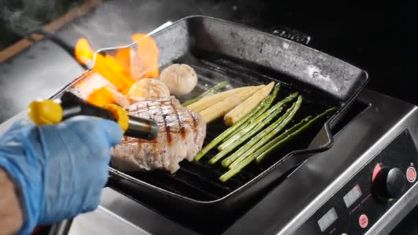 Food video in slow motion. Professional chef in blue gloves cooking with flame on frying pan pork, steak and vegetables with alcohol. hd — Stock Video