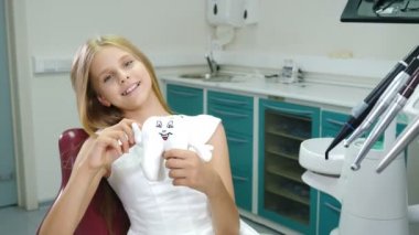 Dişçi muayenehanesinde gülümseyen çocuk. Küçük hasta kameraya bakıyor ve gülümsüyor. Başparmak yukarı. Dişçi koltuğundaki çekici genç kız. Diş koruma konsepti. 4K 'da çekilmiş.