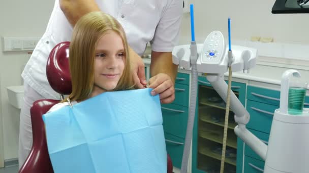 Una niña bonita en la silla dental. Odontología infantil. Dentista preparando poco paciente para el tratamiento. Dentista masculino poniendo una servilleta en un paciente joven. Concepto de atención sanitaria.Caliente en 4k — Vídeo de stock