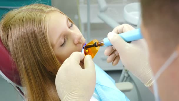 Pequena menina bonito na clínica odontológica recebe tratamento para preencher uma cavidade no dente. Restauração dentária e polimerização de materiais com luz UV. Tiros em 4K — Vídeo de Stock