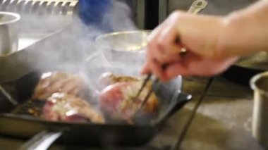 İlk plan - odaklanmamak. Izgarada kızartılmış et. Restoranın mutfağındaki görüntüler. Kapatın. Pişirme maşasıyla aşçıbaşı biftek yapıyor. 4K 'da çekilmiş.