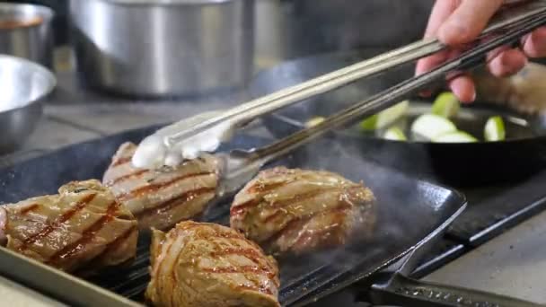 Chefe usando tong para virar carne no grelhador. .. Grelha, bife saboroso. Fecha. Restaurante rush na cozinha. Chef transforma bacon com pinças de metal na superfície quente da grelha. homem com pinças pega, voltas torneiras — Vídeo de Stock