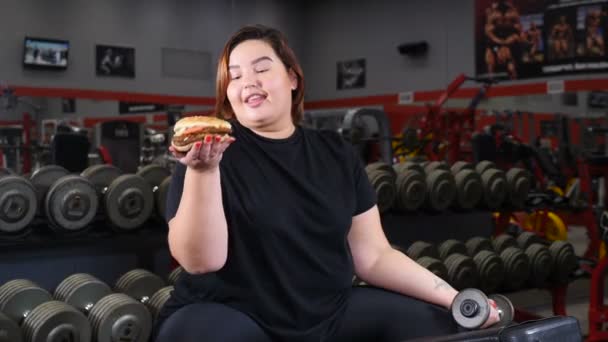 Volontà e perdita di peso concetto. Donna obesa dall'aspetto piacevole che si prende una pausa per lo spuntino durante l'esercizio in palestra. Ragazza mangiare panino enorme guardando la fotocamera. Girato in 4k — Video Stock