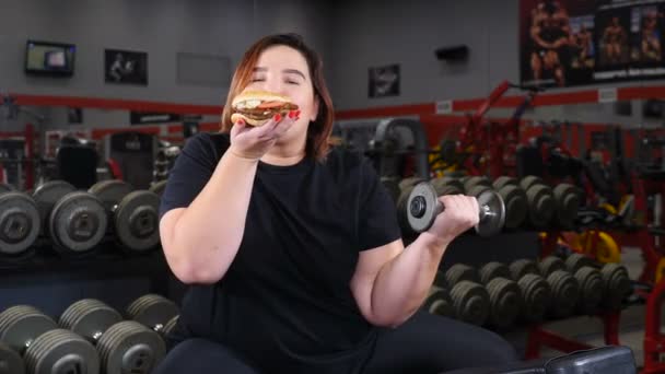 Abnehmen und Willenskraft-Konzept. Dicke Frau im Fitnessstudio bei Übungen im Fitnessstudio. Mädchen entscheiden sich für Junk Food anstelle eines gesunden Lebensstils. Schuss in 4k — Stockvideo