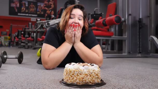 Mujer atractiva y gorda tumbada en el suelo del gimnasio con un pastel enorme. campanas y variedad de máquinas de fitness en segundo plano. concepto anti dieta. Disparo en 4k — Vídeo de stock