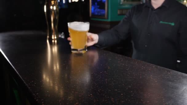 Een close-upshot van een barman die een bierbeker op een donkere bar vasthoudt en rolt. Mok bier wordt geleverd aan mannelijke klant in stijlvolle bar, slow motion. Barman gooit glas met bier. hd — Stockvideo