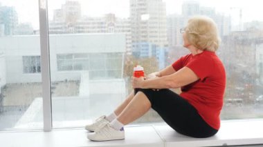 Eğitimden sonra spor salonunun pencere eşiğinde oturan pozitif ve aktif bir kadın. Arka planda panoramik pencereler var. Mutlu ve rahat bir bunaklık. 4K 'da çekilmiş.