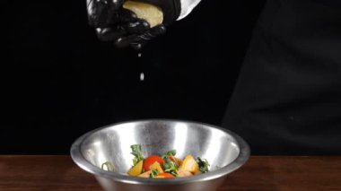 Şefin siyah eldivenli eli, taze limonu taze salatayla çelik kaseye sıkıştırıyor. Siyah arka planda çekilmiş. Yavaş çekimde su sıçrıyor ve düşüyor. Tam Hd