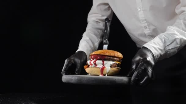 Fast food lokantası konsepti. Şef lezzetli bir burger servis ediyor. Lezzetli bir cheeseburger ve siyah arka planda taze salata malzemeleri. Erkek aşçı yemek sunar. — Stok video