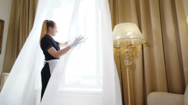 Havalandırma ve havalandırma odası. Üniformalı hizmetçi dantel perdeyi açıyor, otel odasına ışık ve temiz hava giriyor. Otelde temizlik. 4k görüntü