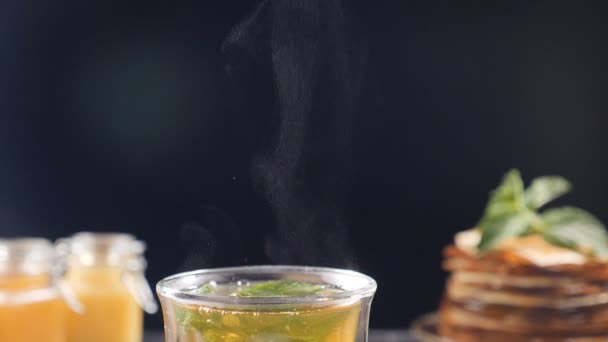 Auf schwarzem Hintergrund dampft aromatisierter Tee, Honig und Pfannkuchen stapeln sich unkonzentriert. Schuss nach vorne. Nahaufnahme. Dampf steigt über Tasse mit Kräutertee auf. Zeitlupe. Volle Konzentration — Stockvideo