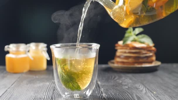 Häll te med apelsin, citron och lime ur genomskinlig tekanna i glas med nint.Brygg te med honung och pannkakor i bakgrunden. Slow motion-te. Fullständig hd — Stockvideo