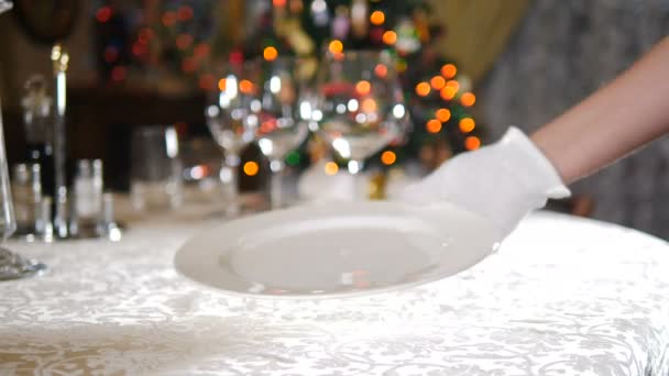 Концепція реставрації. Новий рік і веселий різдвяний стіл. Офіціант у рукавичках кладе серветку на стіл на задньому плані. Світло святкового дерева блимає. страви з окулярами на святкуванні. — стокове відео