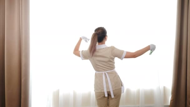 Trabalho doméstico e conceito de limpeza - empregada doméstica feliz em uniforme ou empregada doméstica dançando na frente da janela. Menina muito alegre dançando no quarto de hotel. limpeza da casa e entretenimento. 4 k tiro — Vídeo de Stock