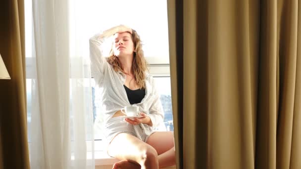 Hermosa joven tierna mujer elegante sentada en el alféizar de la ventana, posando, sonriendo a la cámara. Acogedor apartamento o habitación de hotel. La luz del sol atrás. Disparo en 4 k — Vídeos de Stock